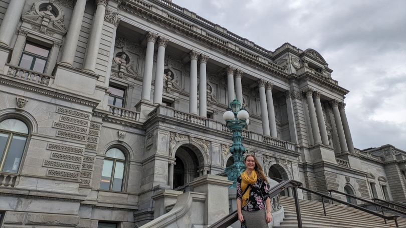 The author, Lily, in front of the Library of Congress in a floral shirt with a yellow scarf. The sky in the background is white and grey.