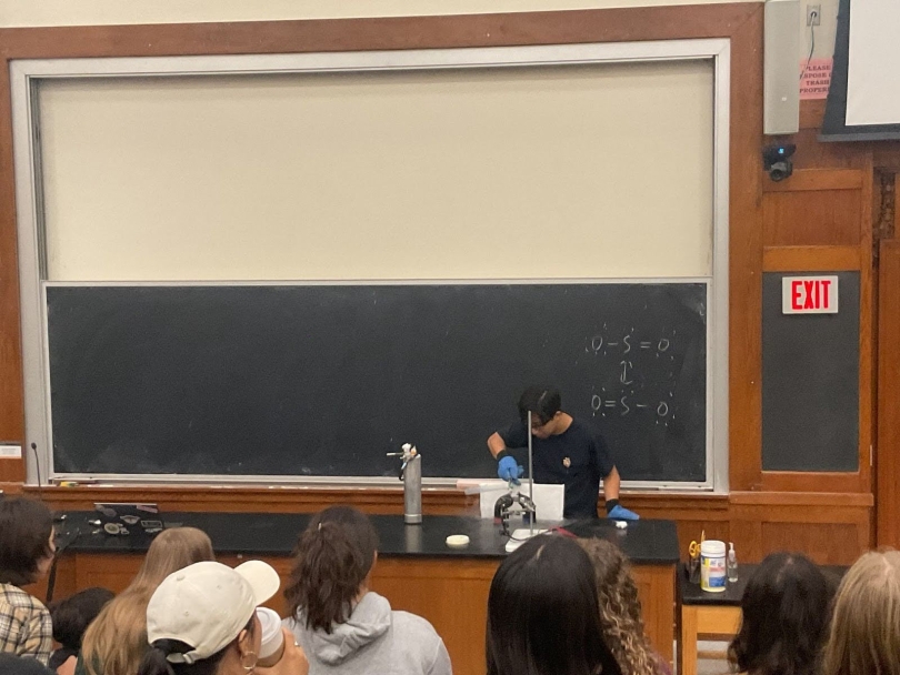 A man standing in front of a chalkboard wearing blue safety gloves and pouring a liquid onto a setup of two metal blocks positioned next to each other. The liquid is so cold that there is vapor coming off of it, and there are students sitting opposite 