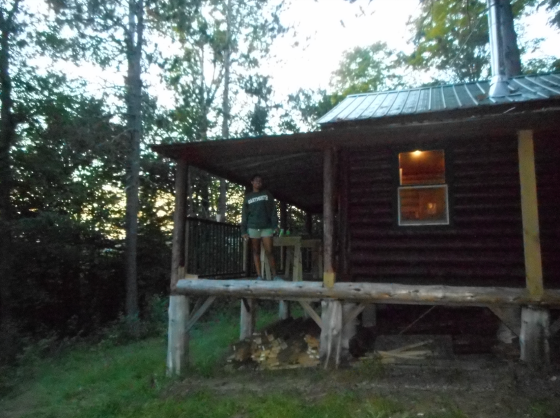 A dark brown cabin with a porch on the left side. A girl is standing on the porch posing for a photo while wearing a green Dartmouth sweatshirt and gray shorts. There is a faint light on in the cabin which you can see from outside. The sky is greyish.