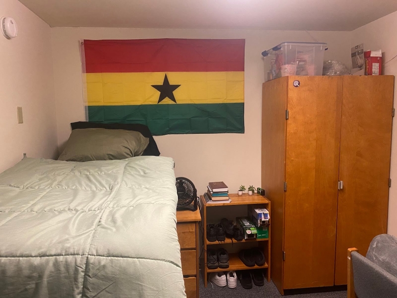 On the left side is a bed laid with a green comforter. On the back wall is a Ghanaian flag. In the far center and right are a shoe rack and a wardrobe respectively. There is a chair in front of the wardrobe. 