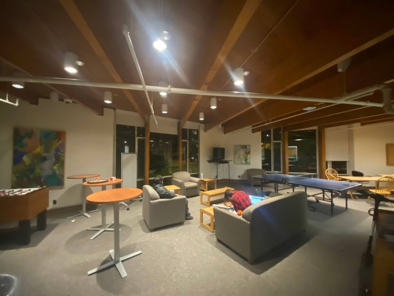 A room with white walls and gray carpeting. In the center of the room are two gray couches with small tables in front of each. On the left side of the room are three taller tables and a foosball table. On the right side of the room is a ping pong table 