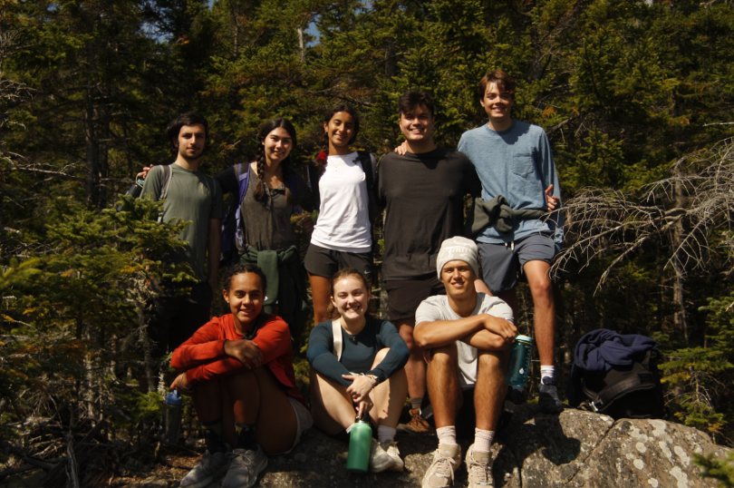 A group of eight people are posing for a photo on a rock in the woods. The trees behind them are full and a dark forest green color. Five people are standing on the rock and three people are sitting down in front of them.
