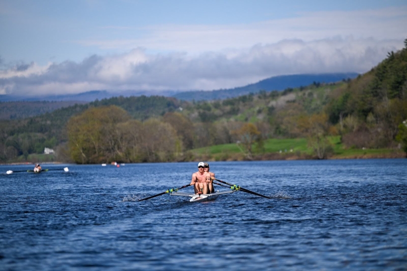 two rowers sit in a boat on a cloudy day on a river with green trees on the shore
