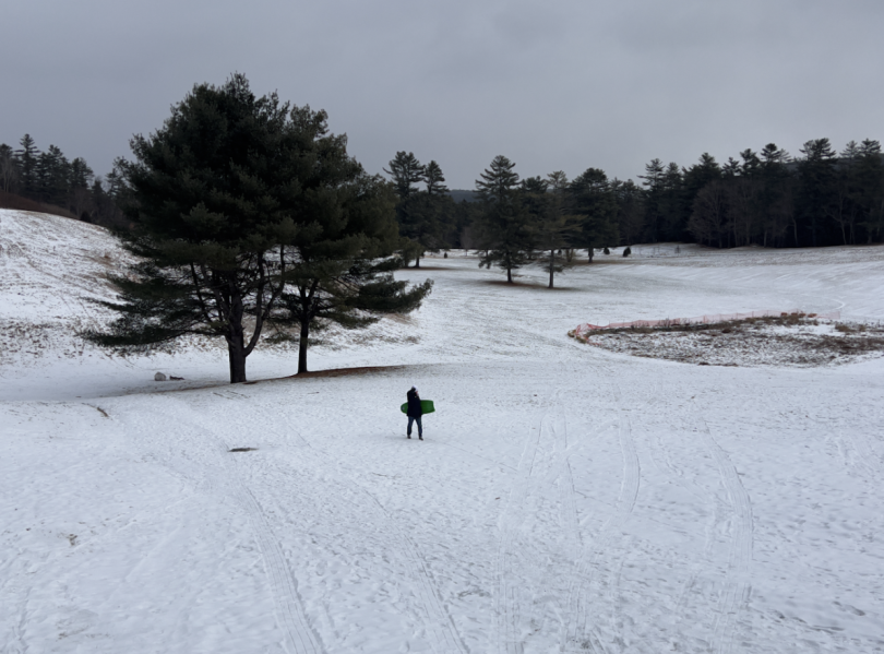 Alt: a white, misty field of slopes with footsteps and trails left behind by sleds. A person stands in the distance with a green sled looking at the camera striking a pose! 