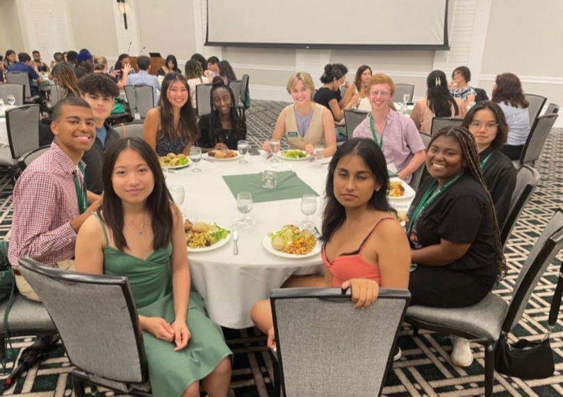 Imagine featuring a fancy dinner inside a room with several tables filled with people can be seen in the background. In the main frame several people sitting around a circular table can be seen posing for a picture. 