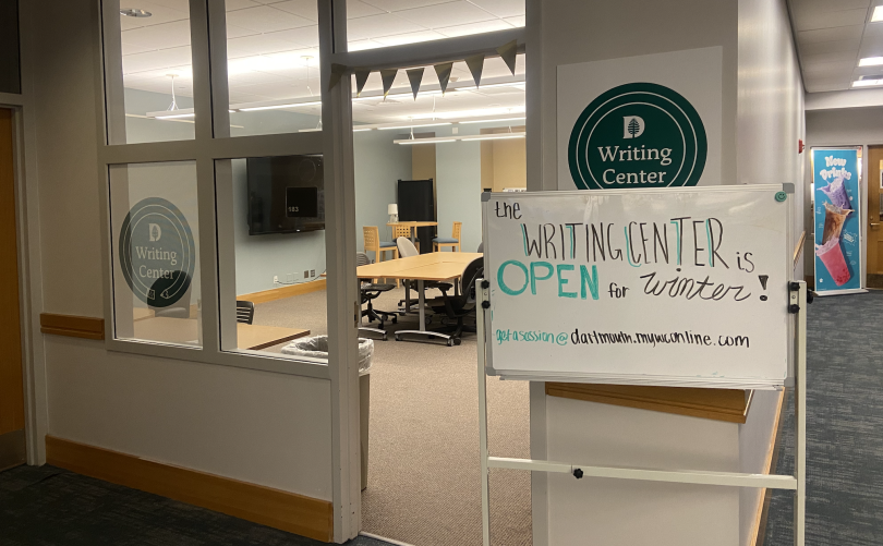 A room with a brown desk in the middle. Just outside the door, there is a whiteboard that reads," Writing Center Open for the Winter." There is also a green circle on the wall with white text inside that reads "writing center."