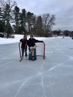 Abbi and a friend in front of a hockey goal. The friend is taking a break from skating by sitting on a trashcan