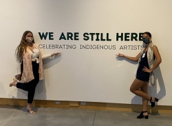 Elizabeth Coleman '21 (left) and Mikaila Ng '22 (right) standing in front of "We Are Still Here" exhibit banner
