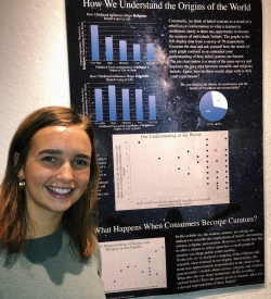 Abbi posing with her poster on display at the Fairbanks. The title of the poster is "How We Understand the Origins of the World"