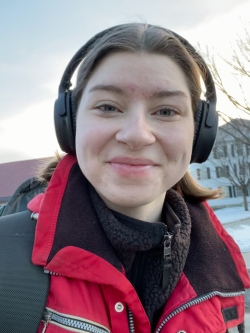 A selfie of me wearing my headphones with a big, red ski jacket on. I am smiling without my teeth, and look tired.