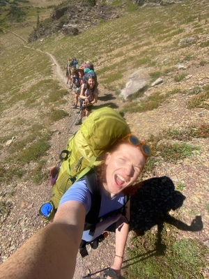 A woman with red hair takes a 0.5 selfie with a group of 7 other students behind her. They are all backpacking. It is sunny out, and they are surrounded by mountains.