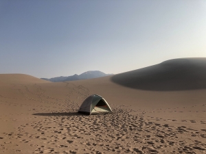 A field of dunes at Great Sand Dunes National Park in Colorado. The visible portion of the dune field is empty except for a tent, with footsteps in the sand leading away from it to the photographer's vantage point.