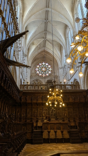 Inside of the cathedral at Burgos with a tall vaulted ceiling and a room lined with carved wooden chairs.