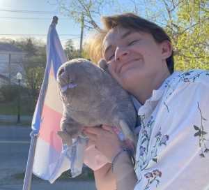 A 19-year-old Dartmouth student in a floral shirt holding a rat stuffed animal in front of a pink, white, and blue transgender pride flag