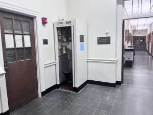 The basement of Dartmouth's Baker-Berry library. To the left is a door, to the right is a hallway, and at the center of the image is a repurposed phone booth with off-white outer walls that match the rest of the hallway. Papers hang on one of its walls.