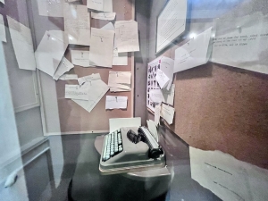 A photo taken through a glass door. On the right is a blue typewriter, and the walls on the back and left are covered in papers with typewritten words on them.