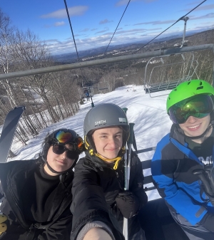 Three people on a ski lift, all smiling and wearing helmets.