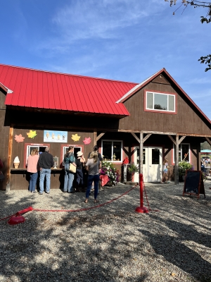 A photo of Mac's Maple, showing the line to get ice cream. There is a brown building with a red roof, and people standing near a pick-up window