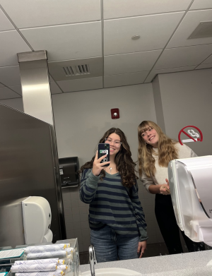 two girls smile at a phone. There is the sink reflected in the mirror and a soap dispenser to the right.