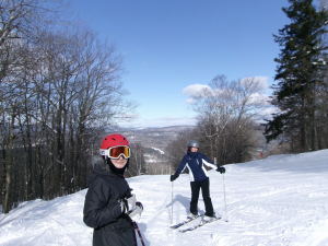 wo women stand on skis and smile at the camera. The sky is bright blue above them and the snow is a bright white beneath their skis.