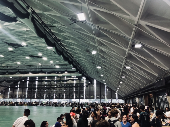 Crowds of students mill about under bright lights while attending a club fair in an indoor track arena. 