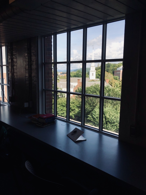 A view of Dartmouth's picturesque campus from an upper-story window of a library study space. Light filters through the window and books lie upturned on tables.