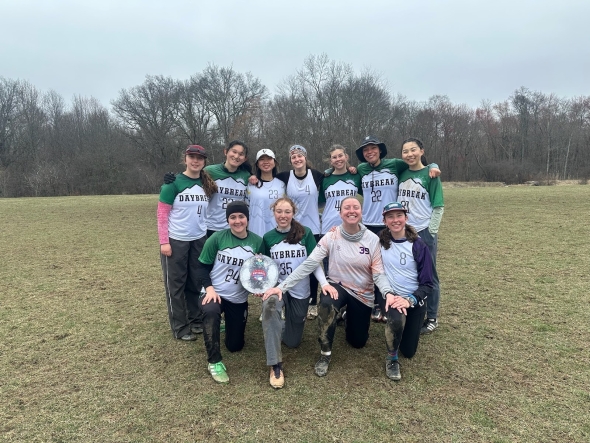 members of the frisbee team stand together and smile at the camera. They are on a green field with leafless trees behind them and the sky is overcast. 