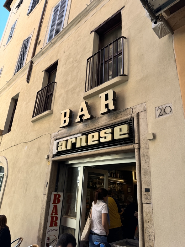 A photo showing the sign and doorway outside of Bar Farnese. There is a woman standing in the doorway of the bar. 