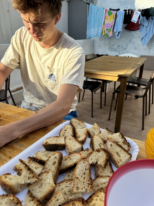 A boy sitting at a table cutting bread into smaller pieces. He is wearing a white shirt and sitting in the sun.