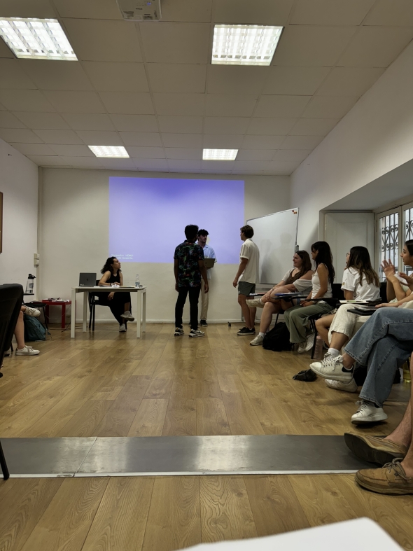 Three students standing at the front of the classroom in front of the project while a seated professor and other students watch them. 