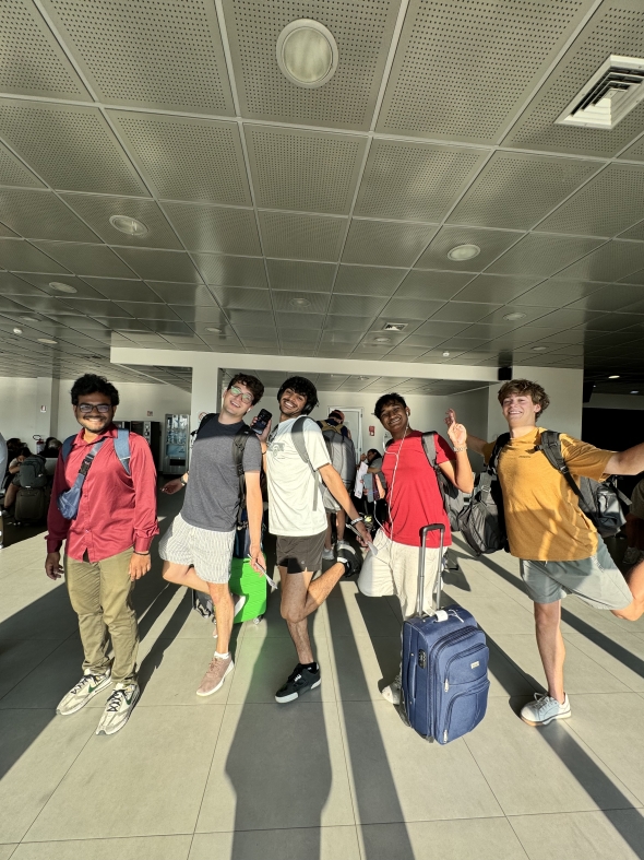 Five boys standing in the airport with their backpacks and carry-on suitcases. They are smiling and each has one leg kicked up in a funny pose.