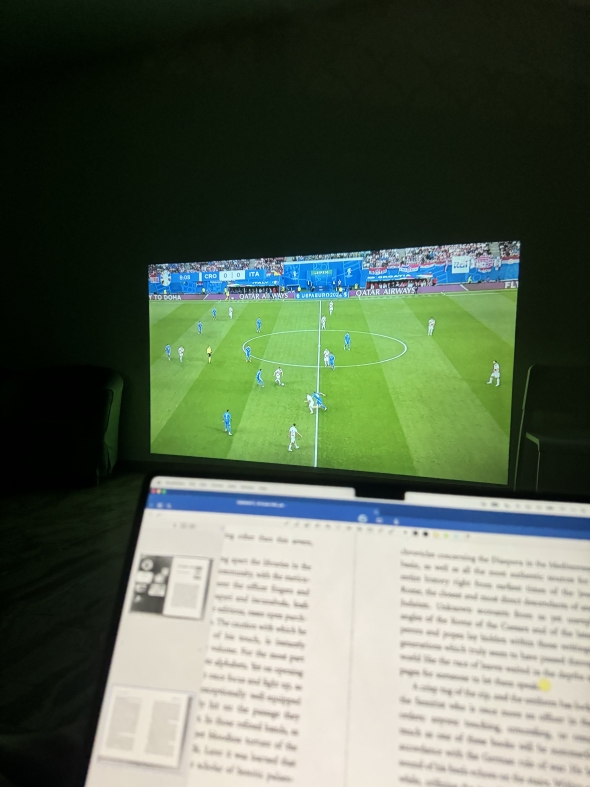 In the foreground, there is a blurred laptop screen. In the background, a projector is showing a soccer match.