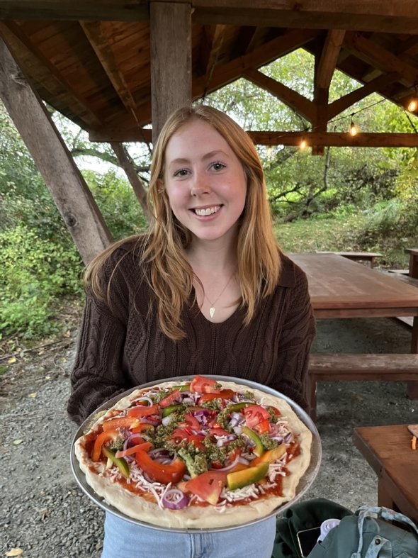 A girl wearing a brown sweater and blue jeans poses with an unbaked pizza in her hands. She is under a wooden structure and there are wooden tables in the background.
