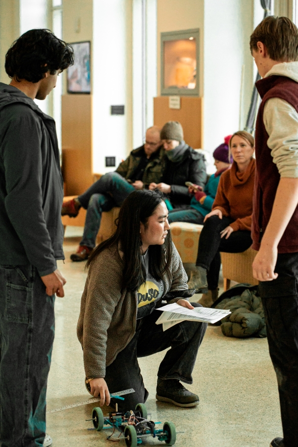 A photo of 2 students standing between 1 volunteer kneeling next to a mechanical car-like contraption.