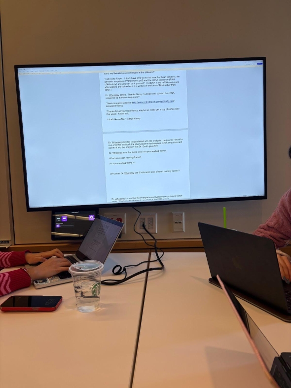 A television with a genetics problem projected. In front of the television is a table with people's laptops on it.