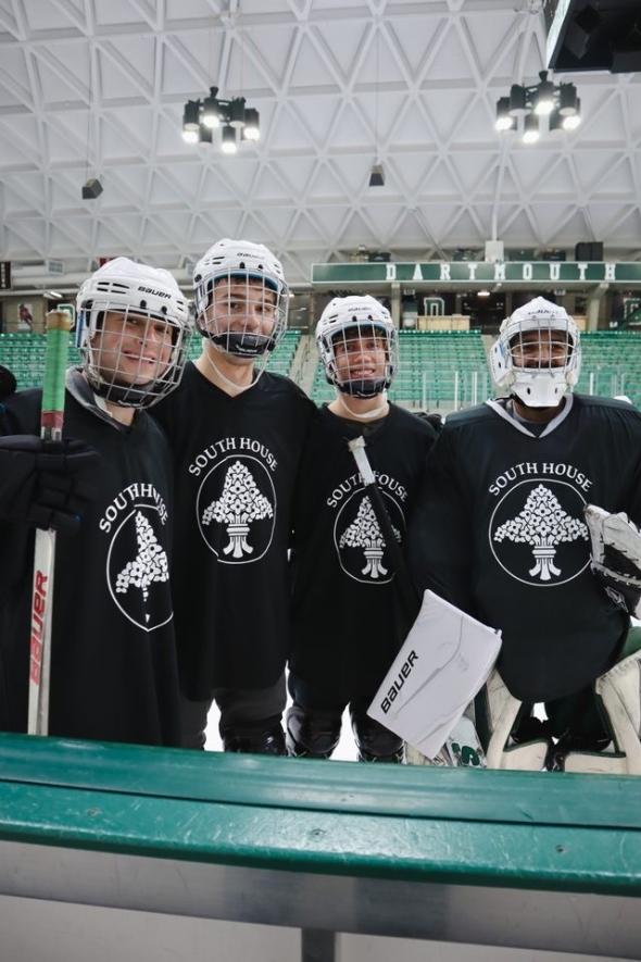 A photo of 4 men standing in a hockey rink, with hockey gear and black shirts saying, "South House".