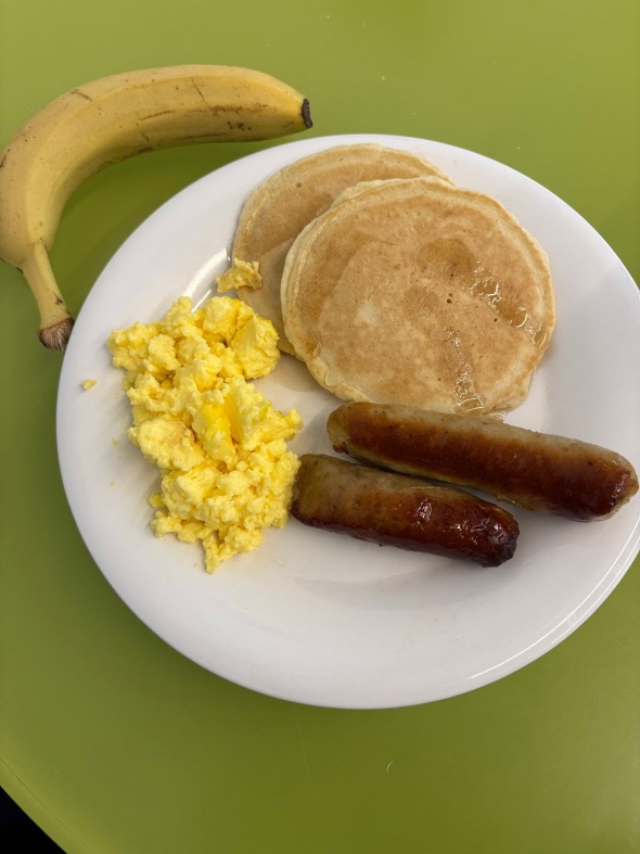 A plate of two pancakes with syrup, two beef sausages and scrambled eggs. There's a banana on the side of the plate as well.