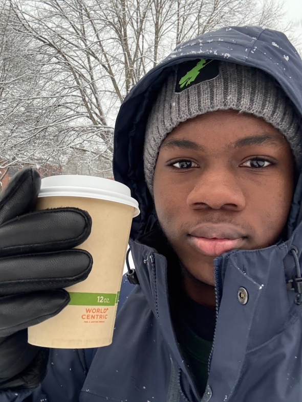 A young man in a winter coat holding a coffee cup. There's a snowy tree in the background.