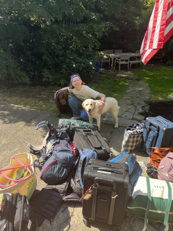 A picture of me smiling next to my dogs, one of whom is a white Labrador and another who is a chocolate Labrador. There is luggage in the foreground of the photo.