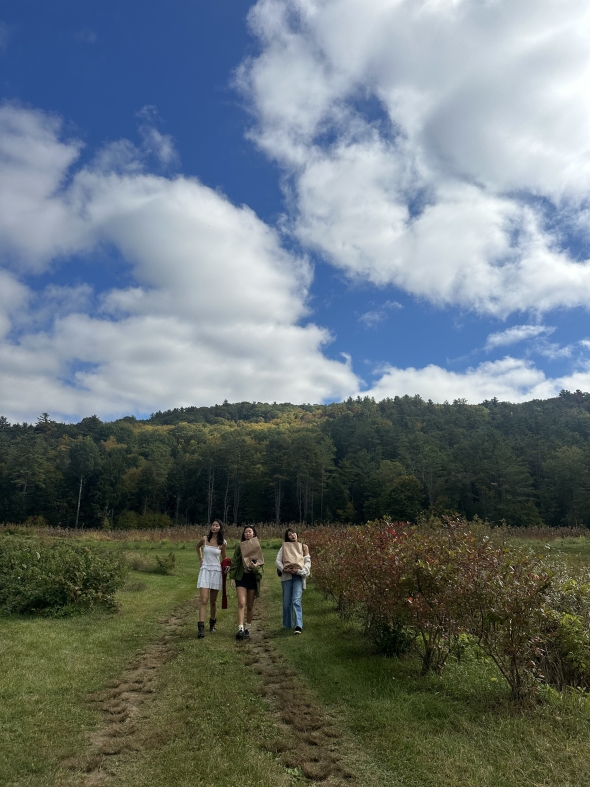 "Three girls walk forward in the distance across a grassy field, with tall green mountains in the background, their slopes dotted with yellow autumn leaves under a clear bluesky.