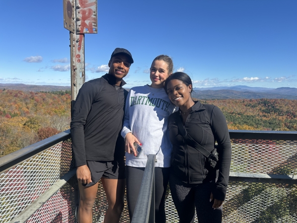 Three friends are smiling, dressed in warm hiking-wear as their backs are turned to a view of glistening fall foliage and colorful trees adorned by a bright, blue sky.