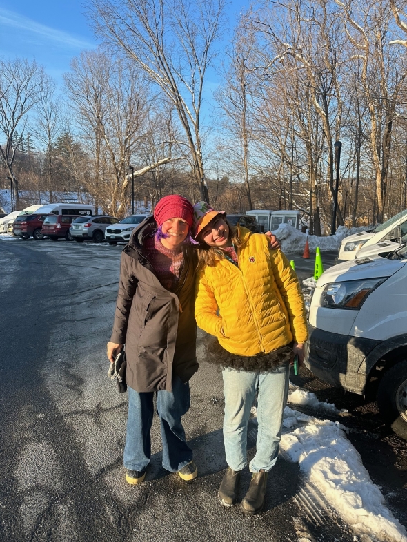 Two women smile next to each other in a parking lot. One is wearing a yellow coat, a tie-dye bucket hat, and a black tutu with jeans. The other is wearing a black coat, a red hat, a purple wig, and jeans. They are wearing matching purple sweaters.