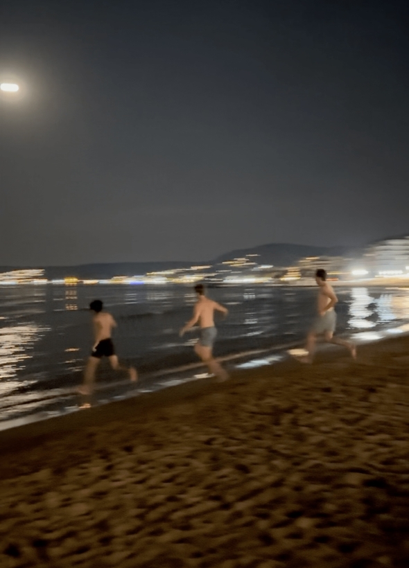 Three boys on the beach running into the ocean. It is nighttime and there is a full moon.