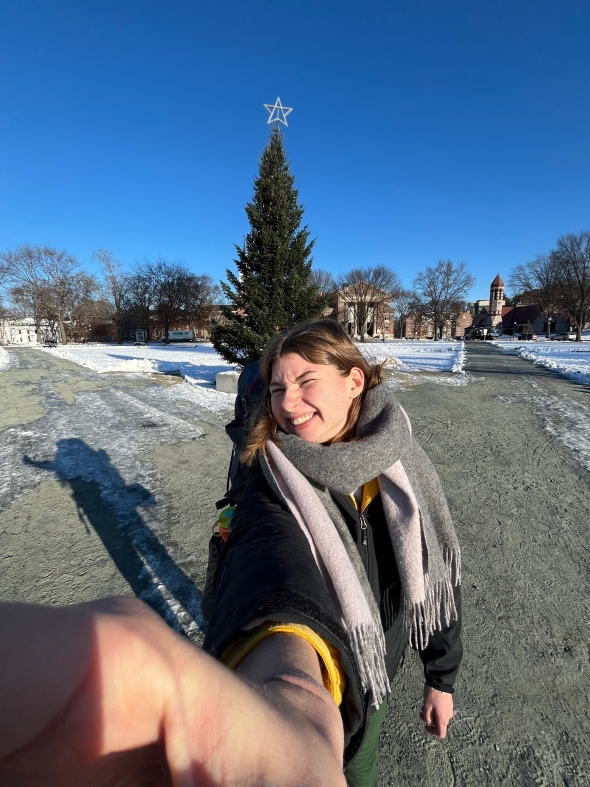 A girl smiles for a 0.5 selfie in the middle of the Dartmouth Green. There is a large pine tree behind her, with a star on top. The girl is wearing many jackets and a scarf.