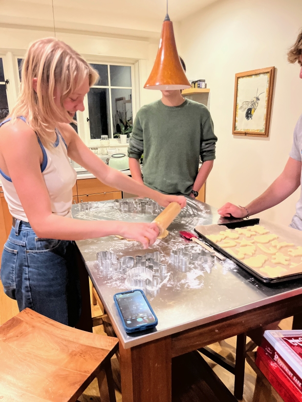 A girl rolls out sugar cookie dough in a nondescript kitchen. She is laughing. There are two other people in the photo, also working on baking cookies.