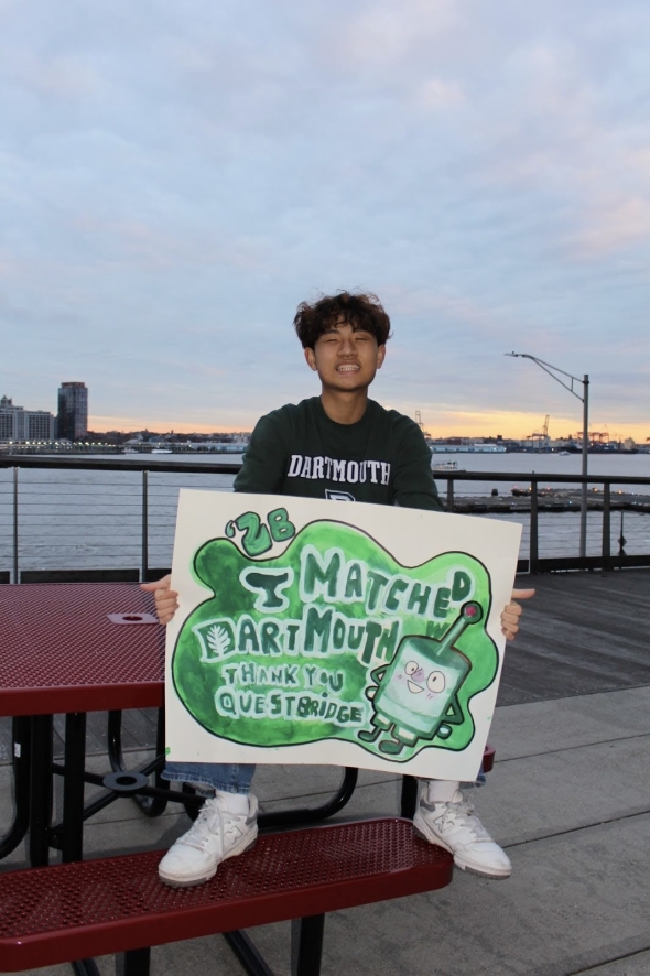 Me sitting on a red bench by the Hudson River in New York, fenced by railings, and soaked in sunset. I am holding up a white poster with a huge smile, stating "I matched to Dartmouth. Thank You QuestBridge"
