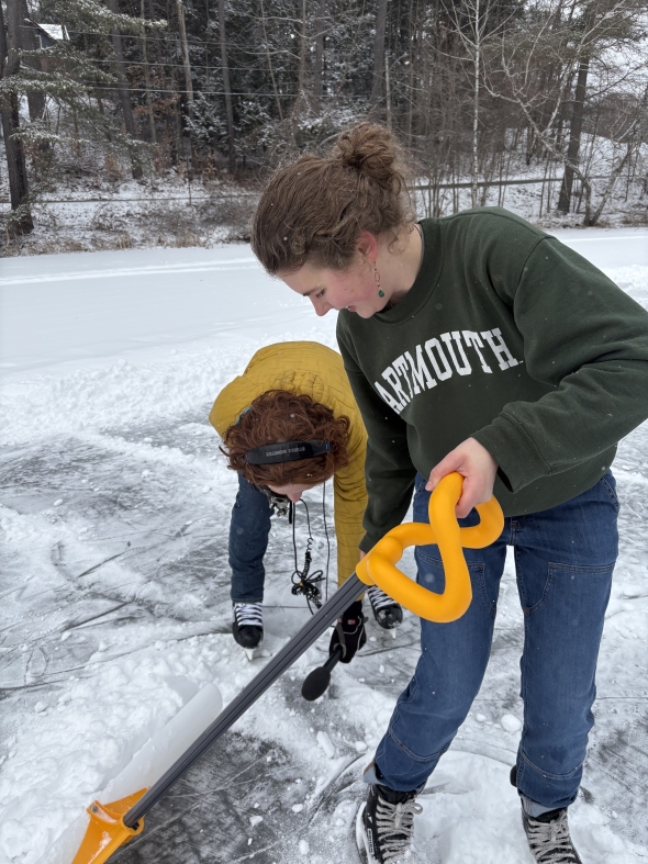 Two women on skates lean over to look at a microphone near the ground. One is wearing a Dartmouth sweatshirt and carrying a shovel, and the other is wearing a yellow coat and is carrying audio equipment.