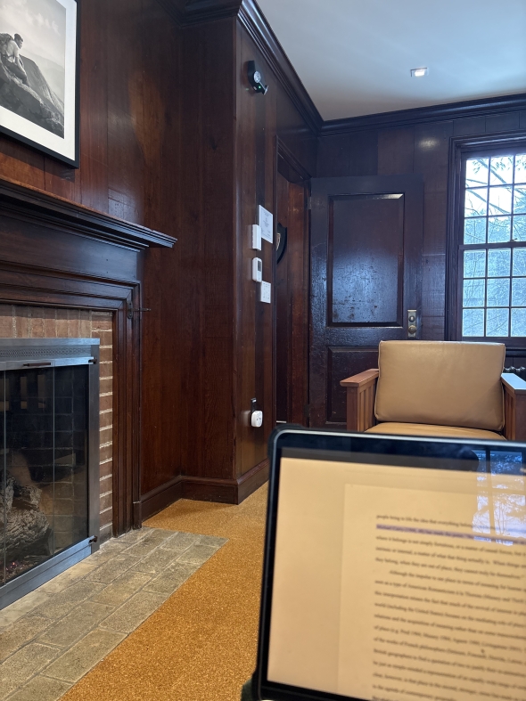 A picture of a computer with a paper pulled up. The background features an armchair and a fireplace. The room is made of dark, red-ish wood.
