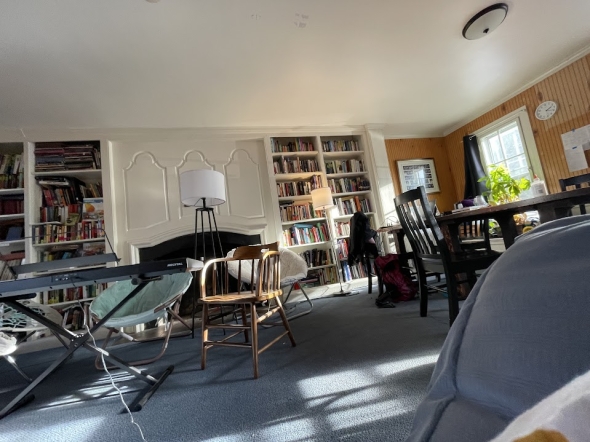 a picture of the Alpha Theta library, visible are a table, a couple of chairs, and an electric piano in front of a white wall and stacks of books
