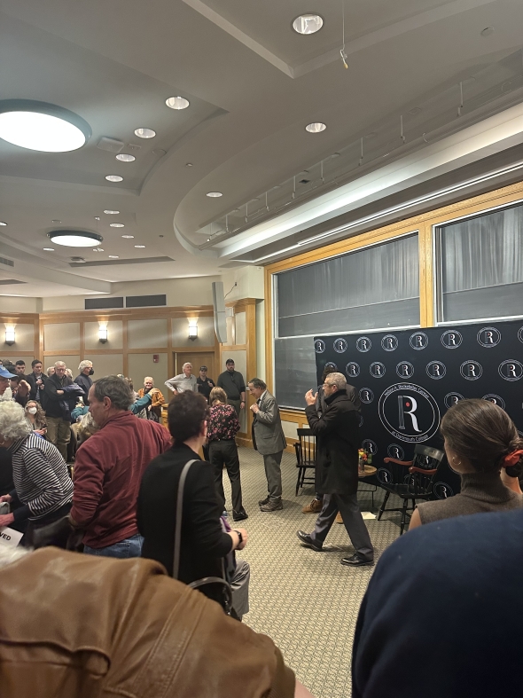 A Rockefeller Center conference room filled with a standing audience gathered around as main speaker Jamie Raskin addresses the crowd from the front of the room.